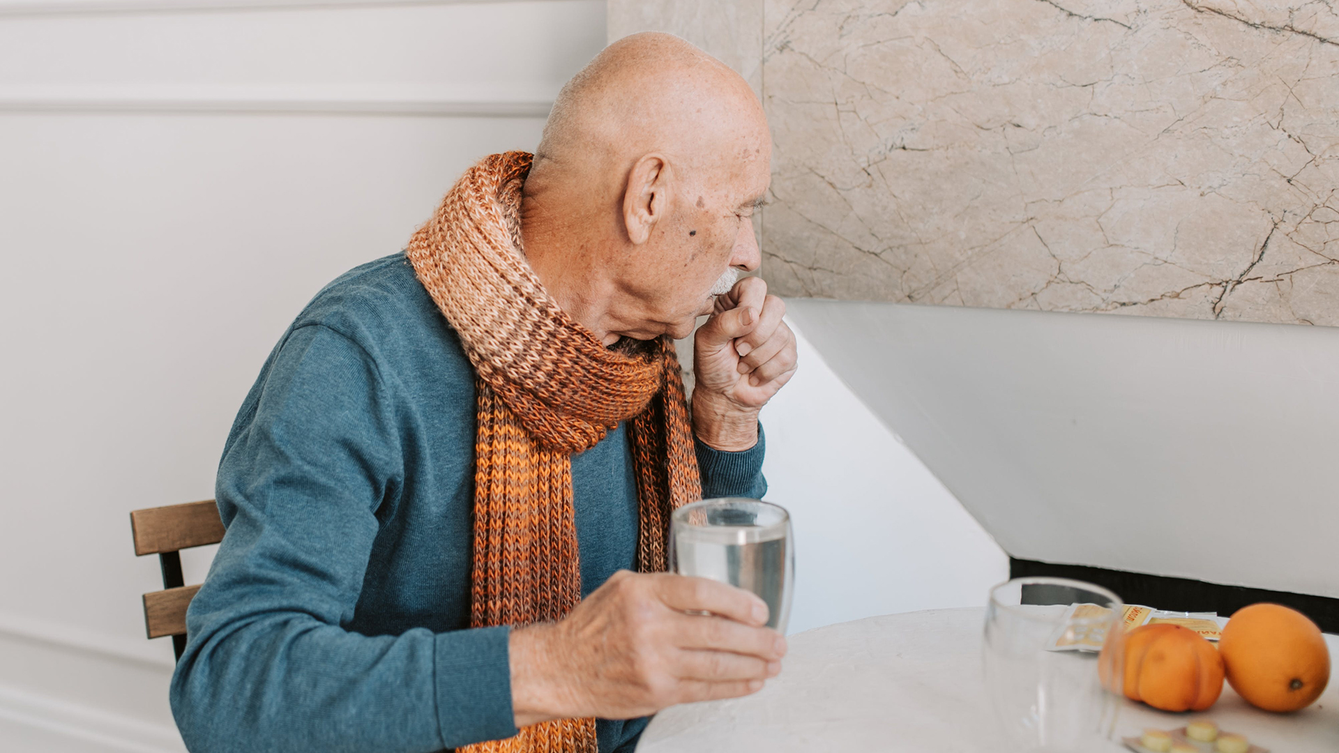 Elderly man wearing a scarf, coughing into his hand and holding a glass of water